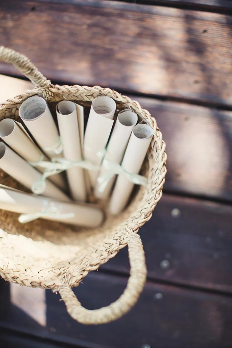 Panier en osier rempli de programmes de mariage roulés et noués de rubans blancs sur un fond de bois sombre.