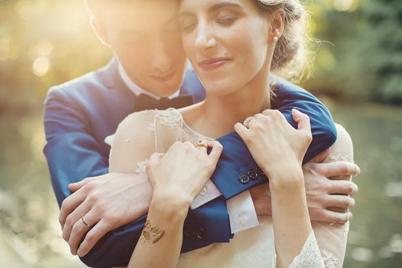 Jeune couple marié s'enlaçant tendrement, lui en costume bleu et elle en robe blanche, dans une ambiance lumineuse.