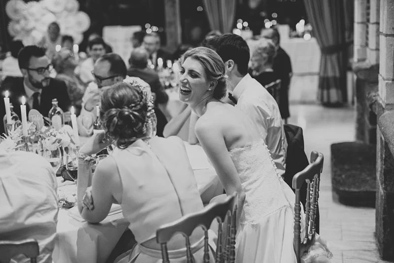 Photo en noir et blanc d'une mariée riant aux éclats lors d'un dîner de mariage entourée de ses invités.