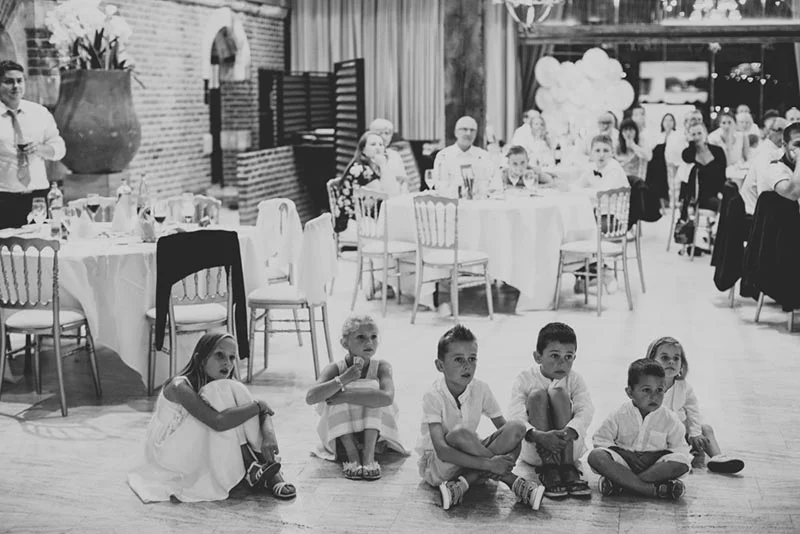 En noir et blanc, quatre enfants assis par terre au premier plan lors du dîner du mariage de Charlotte et Julien.