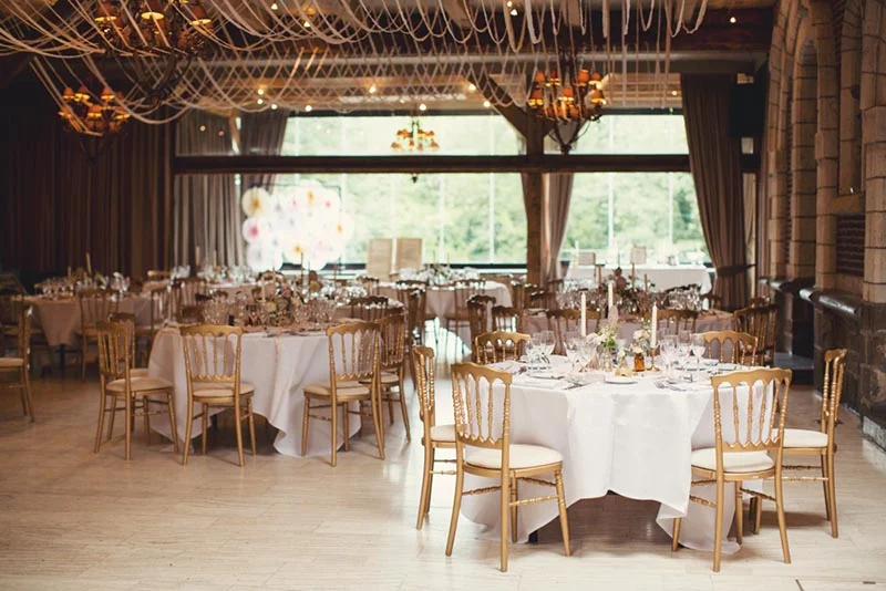 Salle de réception élégante avec tables nappées de blanc, chaises dorées et lustres suspendus sous un plafond drapé.