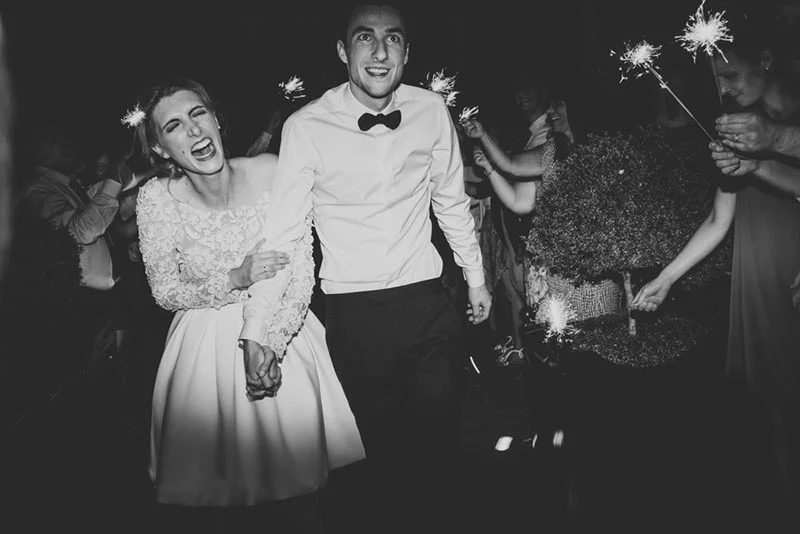 Couple marié souriant marchant sous une haie de cierges magiques, photographie en noir et blanc.