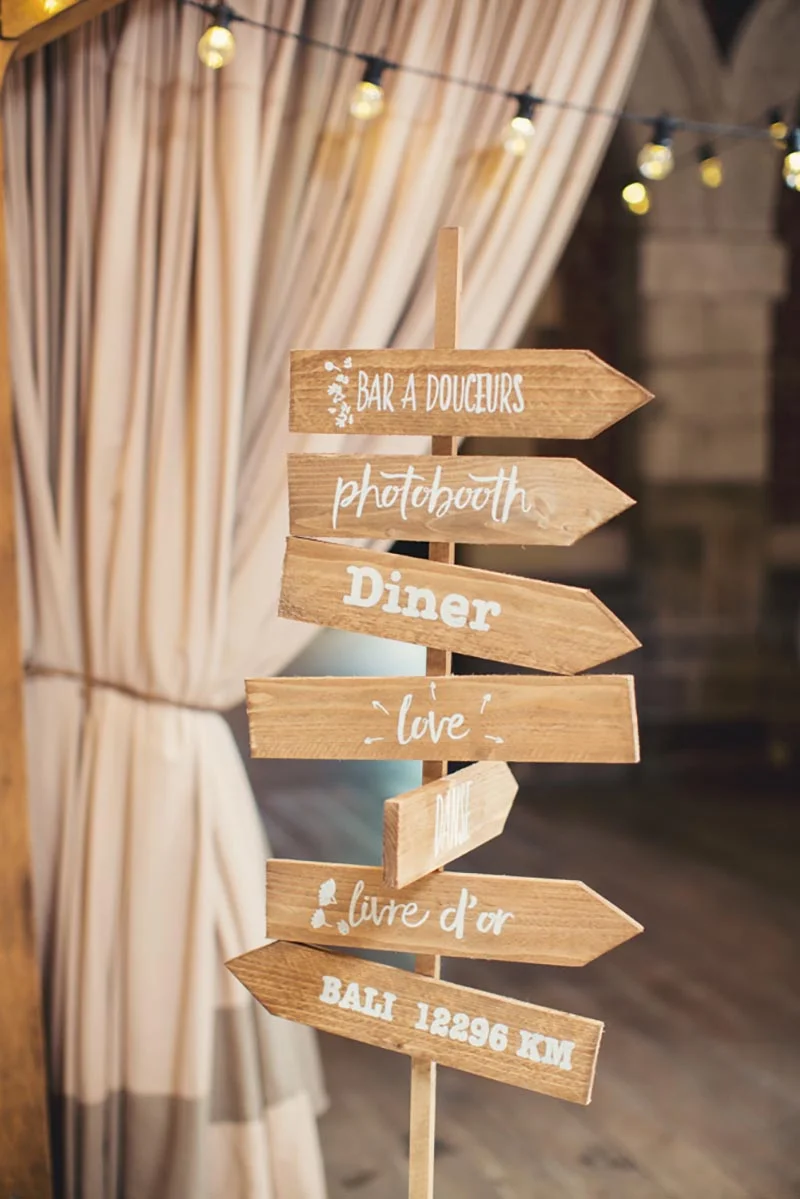 Panneau directionnel en bois rustique indiquant le bar, le photobooth et le dîner devant des rideaux beiges lumineux.