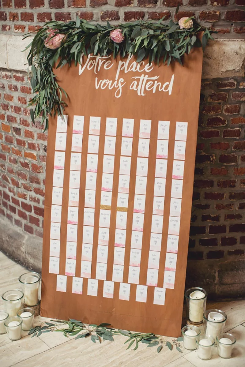 Plan de table rustique en bois brun avec calligraphie blanche, décoré de feuillage et de fleurs roses.