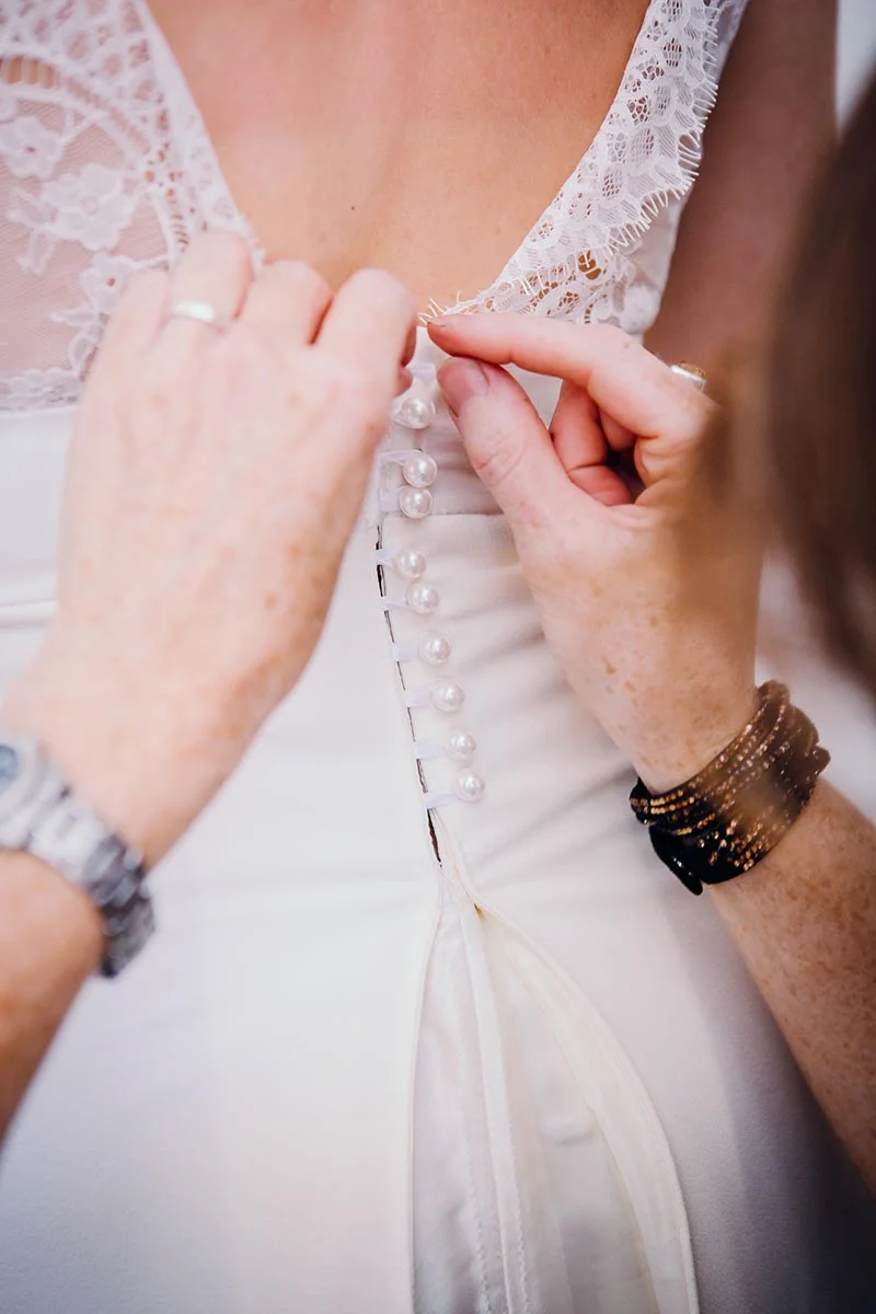 Gros plan sur des mains boutonnant le dos d'une robe de mariée blanche en dentelle et satin.