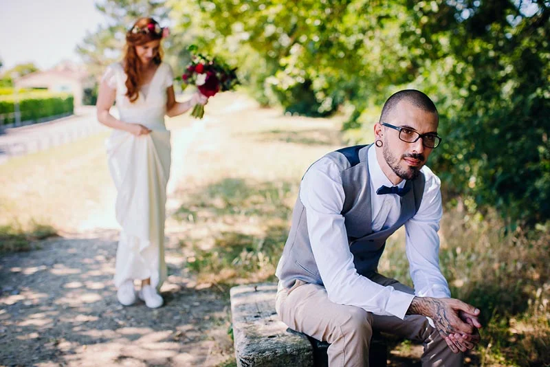 Un marié en gilet gris attend assis tandis que sa mariée en robe blanche s'approche avec un bouquet rouge.