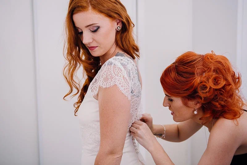 Une femme rousse aide la mariée à ajuster sa robe blanche en dentelle dans une ambiance douce et lumineuse.