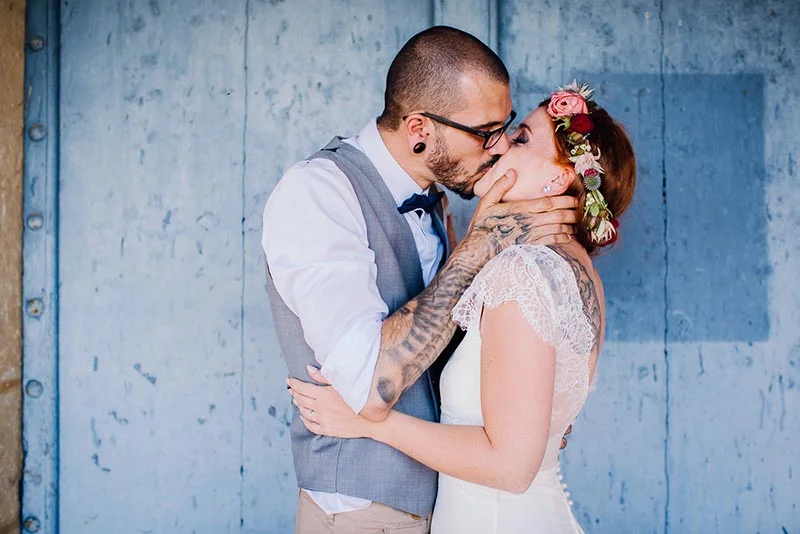 Un couple marié s'embrasse tendrement devant un mur bleu, la mariée portant une couronne de fleurs colorées.