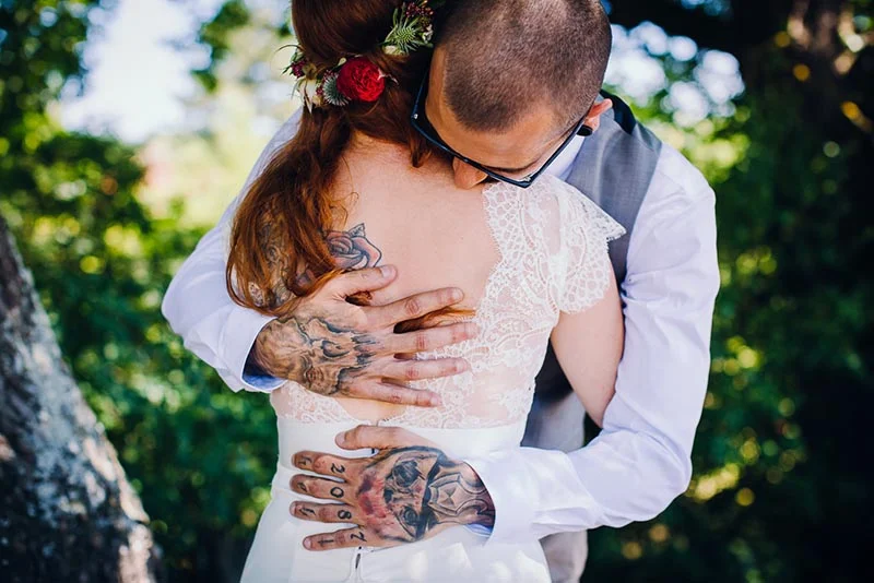 Un marié tatoué enlace tendrement sa mariée en robe de dentelle blanche dans un cadre naturel et lumineux.