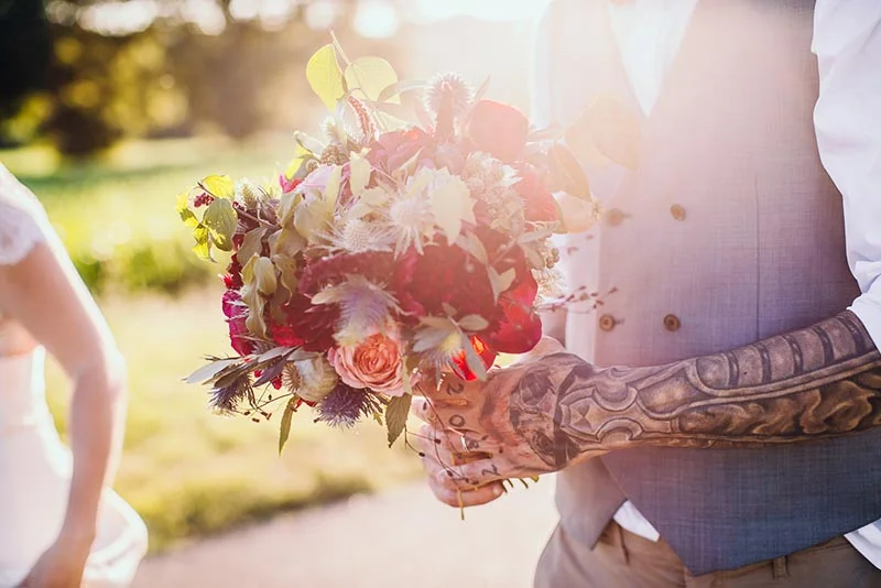 Gros plan d'un marié tatoué tenant un bouquet champêtre rouge et blanc lors d'un mariage en extérieur et lumineux.