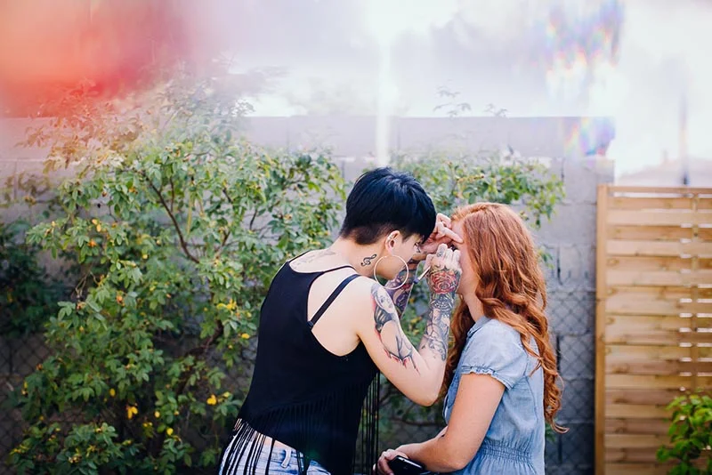 Une maquilleuse tatouée prépare une mariée aux longs cheveux roux dans un jardin lumineux.