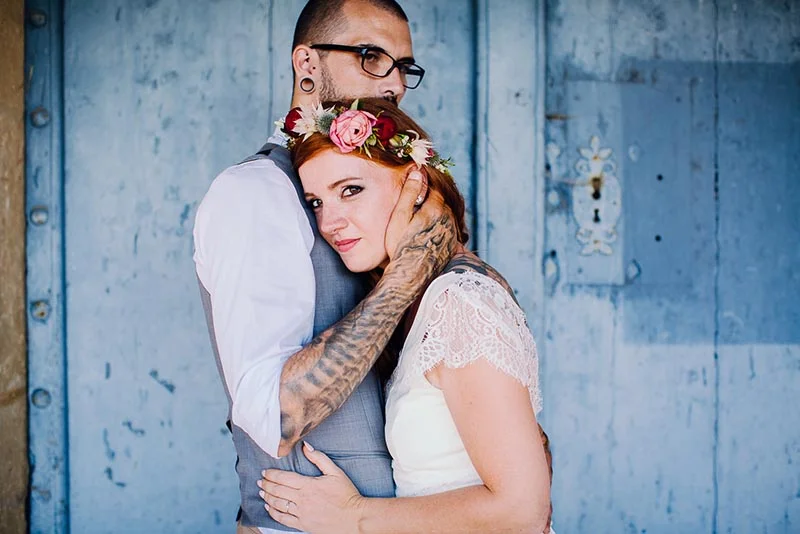 Couple marié s'enlaçant tendrement devant un mur bleu, la mariée portant une couronne de fleurs et une robe en dentelle.