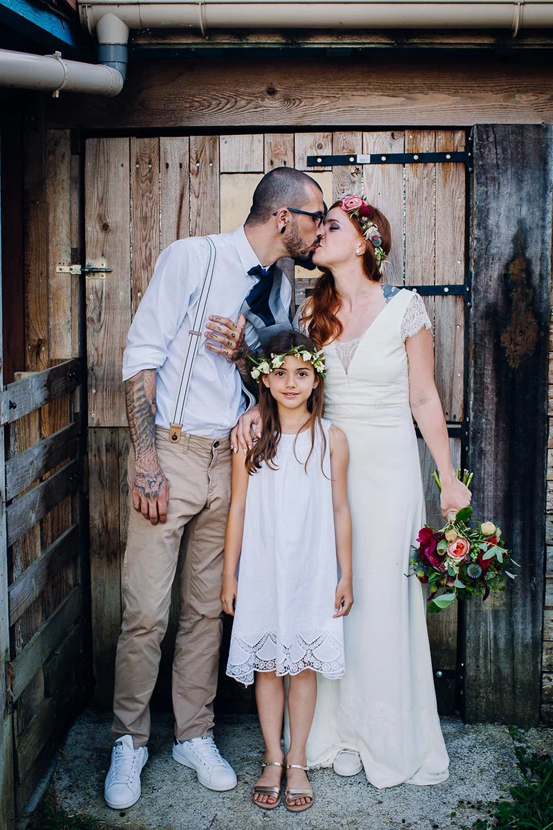 Un couple de mariés s'embrassant devant une porte en bois, accompagnés d'une petite fille en robe blanche.