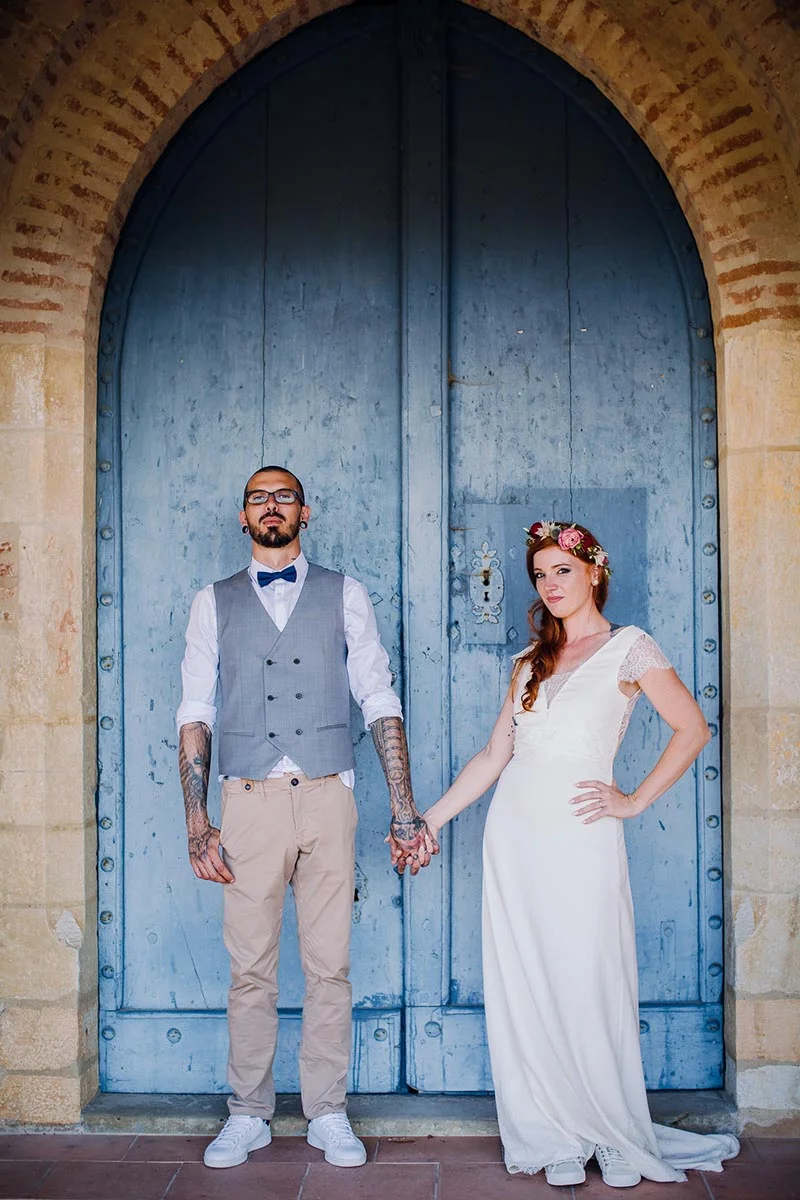 Couple marié posant main dans la main devant une grande porte bleue vintage, ambiance bohème et romantique.