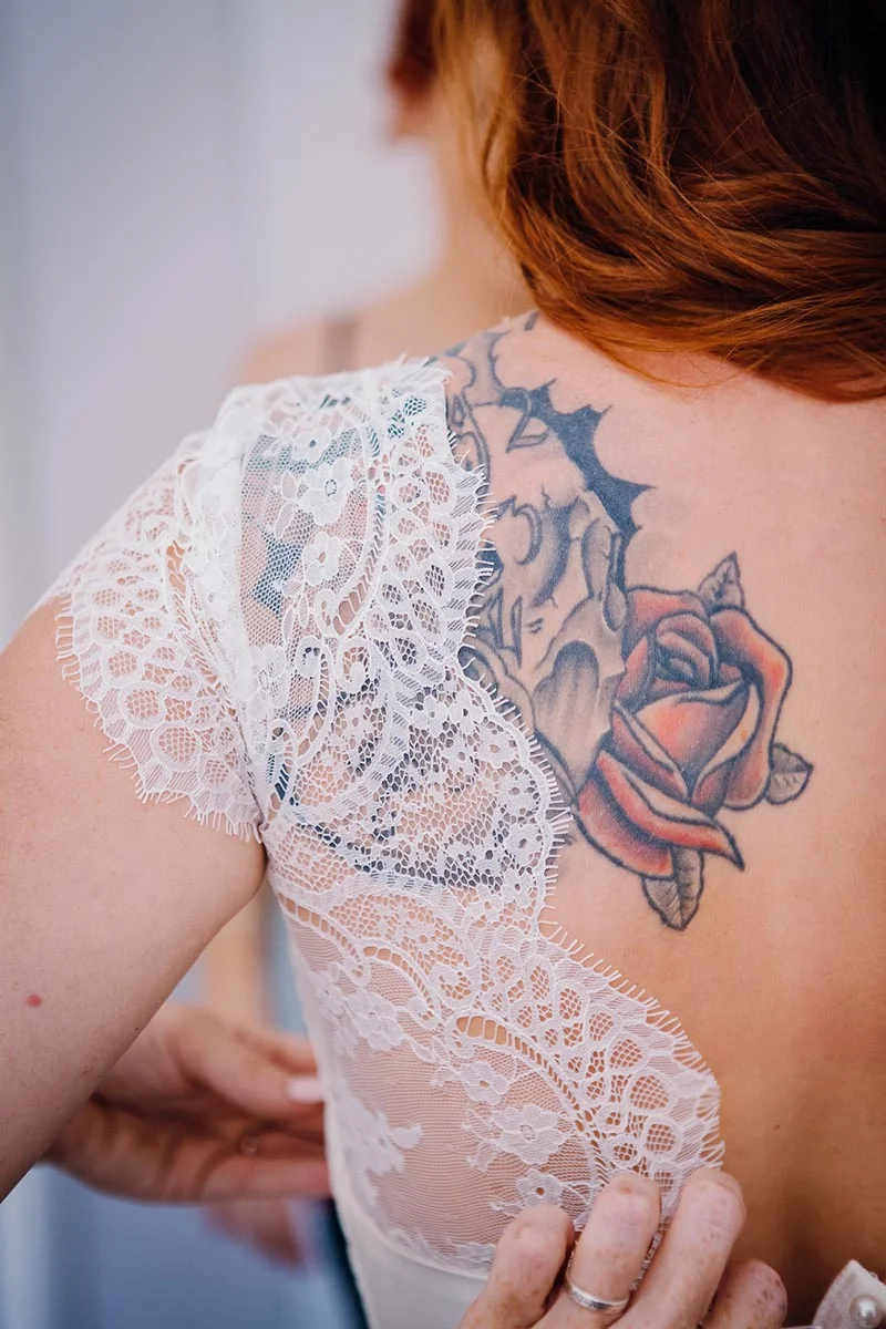Gros plan sur le dos d'une mariée aux cheveux roux, portant une robe en dentelle blanche révélant un tatouage coloré.