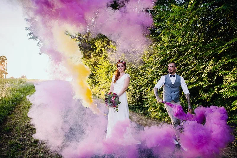Jeunes mariés souriants entourés de nuages de fumée colorée rose, violette et jaune dans un cadre naturel verdoyant.