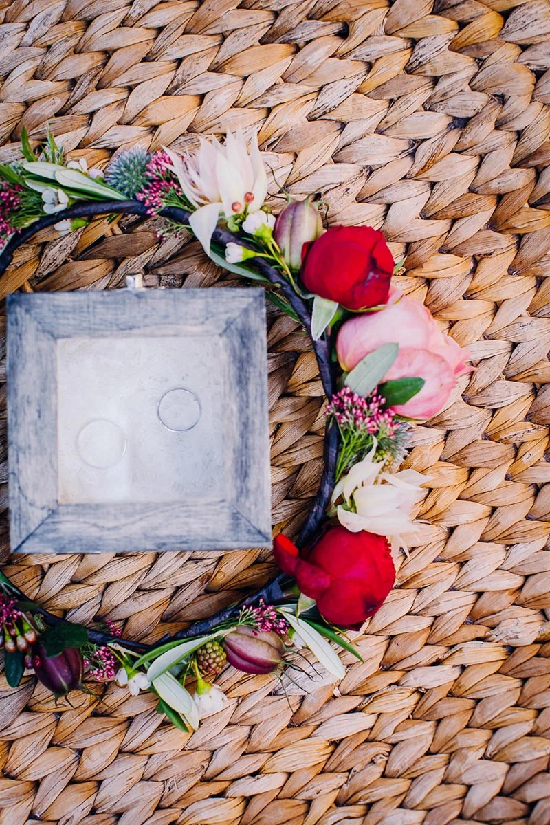 Couronne de fleurs colorées entourant un coffret à alliances en bois sur un tapis en osier tressé.