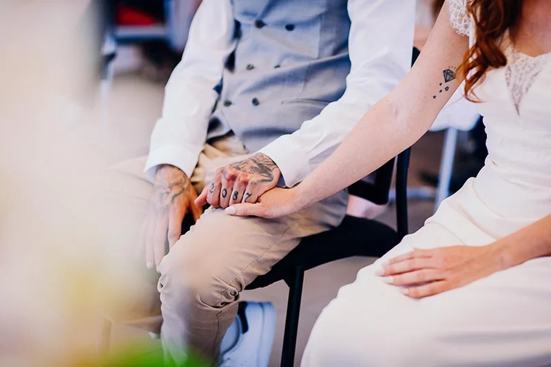 Gros plan sur les mains entrelacées de Virginie et Stéphane, vêtus de leurs tenues de mariage blanches et beiges.