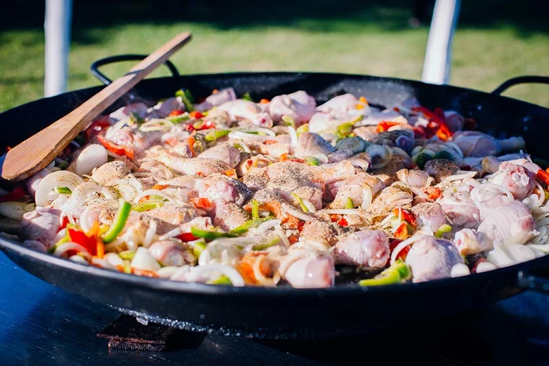 Une grande paella colorée aux morceaux de poulet et légumes cuisant dans un plat noir en plein air.