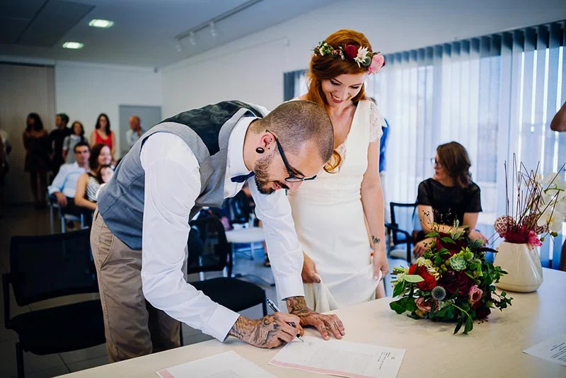 Un marié tatoué signe un document tandis que sa mariée, coiffée d'une couronne de fleurs, le regarde avec tendresse.