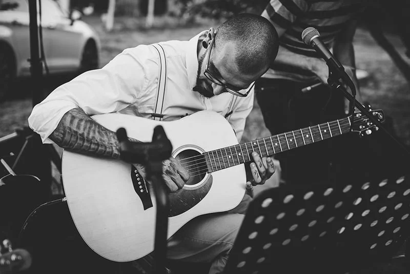 Musicien tatoué jouant de la guitare acoustique lors d'un mariage, cliché en noir et blanc.
