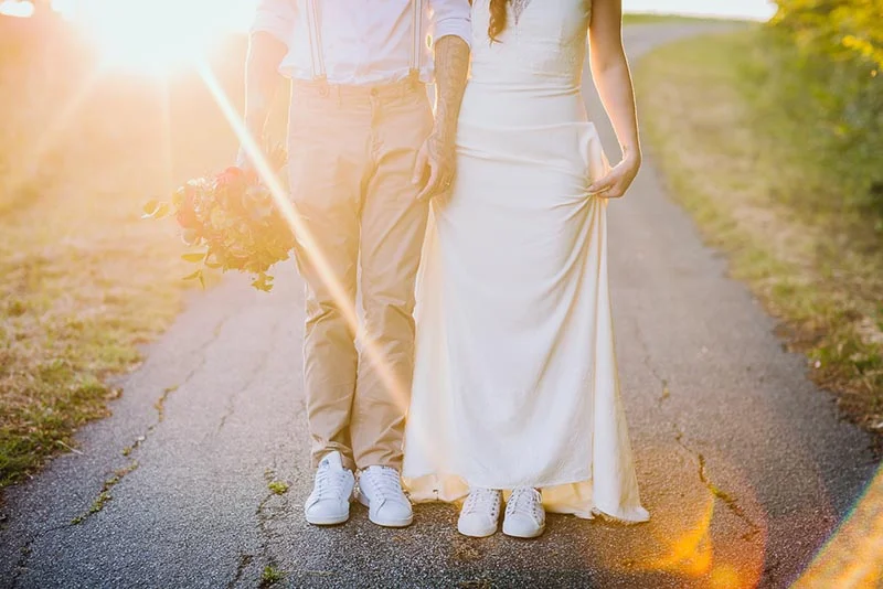 Jeunes mariés en tenue décontractée et baskets marchant main dans la main sur un chemin au coucher du soleil.