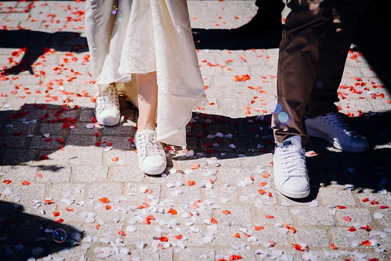 Pieds d'un couple en baskets blanches marchant sur un sol pavé jonché de pétales de fleurs rouges et blancs.