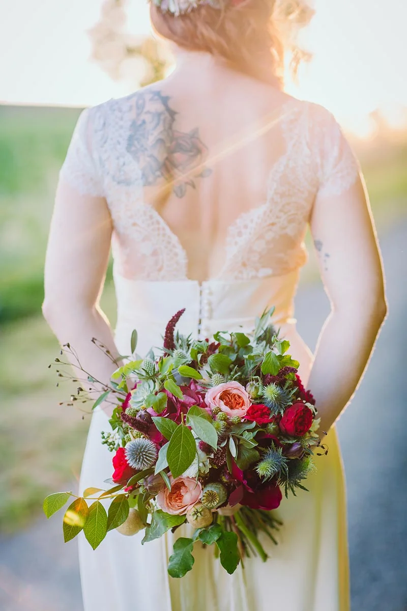 Mariée de dos en robe blanche dentelée, tenant un bouquet coloré de fleurs rouges et roses sous une lumière dorée.