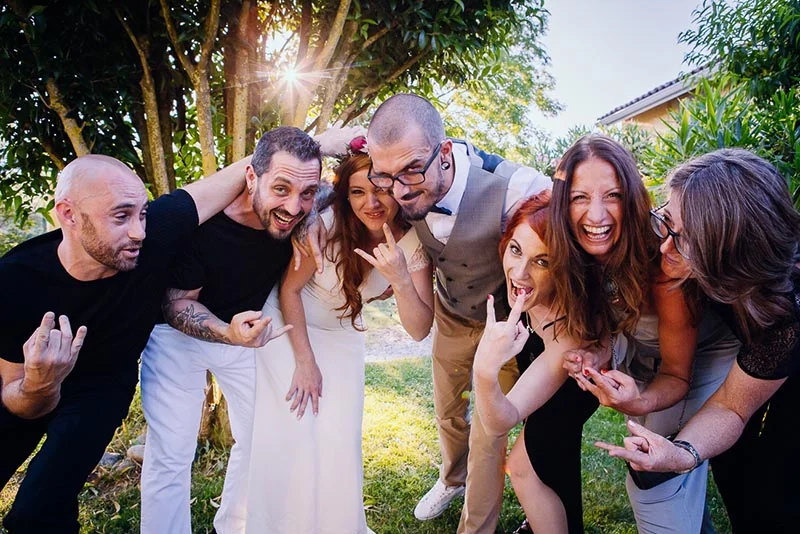 Un groupe d'amis joyeux et décontractés pose avec humour autour des mariés dans un jardin ensoleillé.