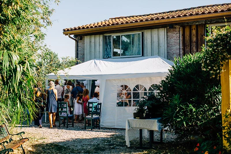 Invités discutant sous un chapiteau blanc devant une maison rustique lors du mariage de Virginie et Stéphane.