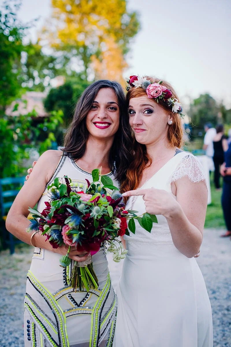 Deux femmes souriantes posent ensemble, l'une tenant un bouquet coloré et l'autre portant une couronne de fleurs.