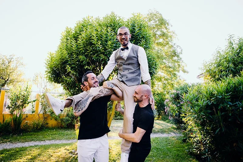 Trois hommes souriants s'amusent en extérieur, l'un porté à bout de bras par les deux autres dans une ambiance festive.