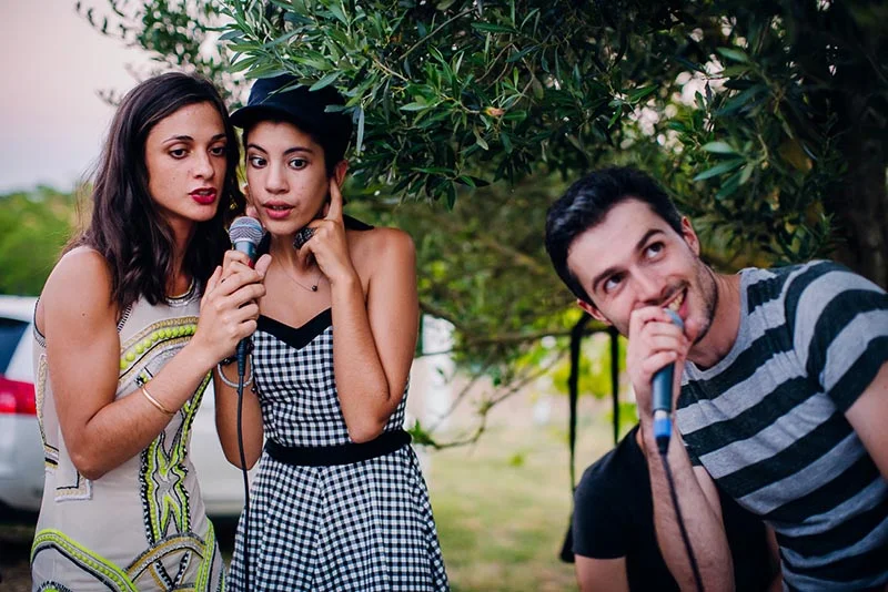 Trois jeunes gens rient et chantent avec des micros sous un olivier lors du mariage de Virginie et Stéphane.