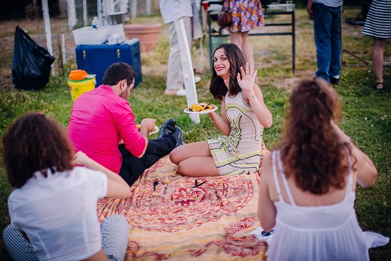 Des invités discutent et partagent un buffet convivial sur une nappe colorée posée dans l'herbe lors d'un mariage.