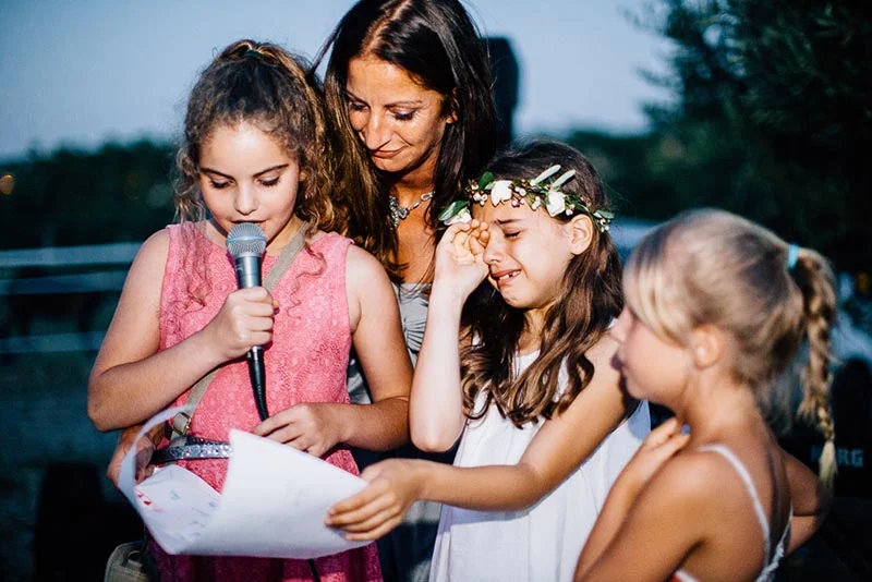 Une femme et trois fillettes émues lors d'un discours, dont une petite fille en robe rose lisant un texte au micro.