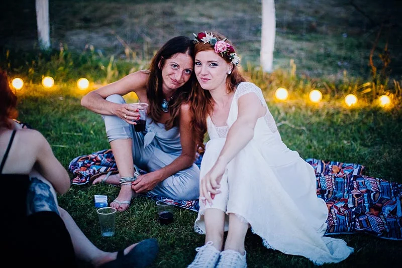 Deux femmes souriantes, dont l'une porte une couronne de fleurs et une robe blanche, se détendent sur une couverture.