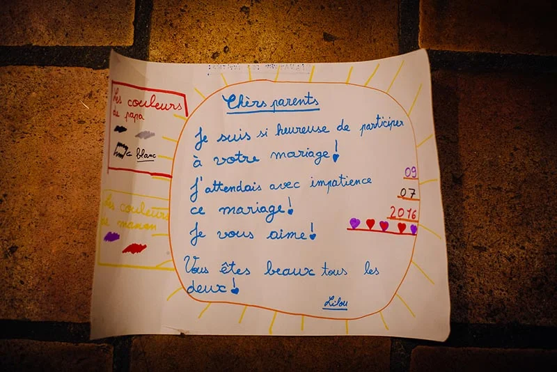 Lettre d'enfant manuscrite et colorée adressée à ses parents pour leur mariage, posée sur un sol en briques.