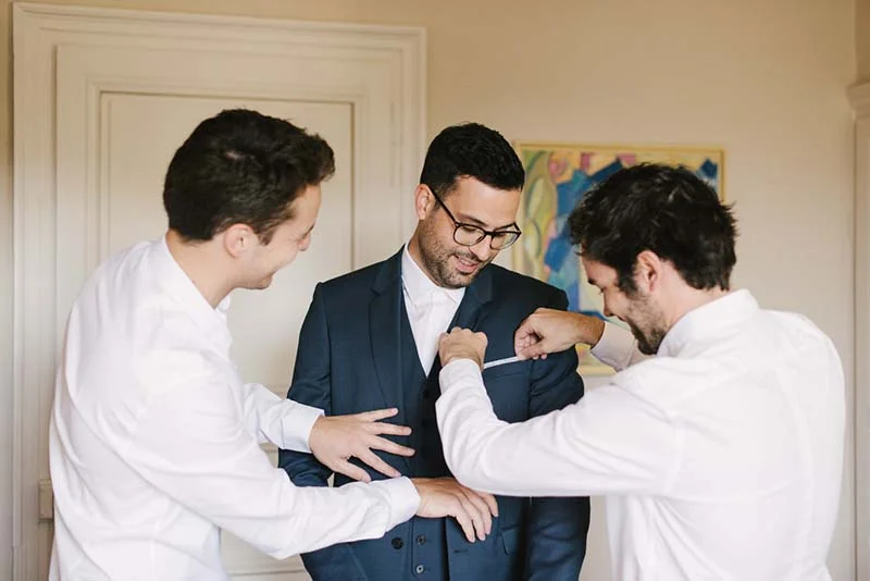 Deux témoins en chemise blanche aident le marié en costume bleu à ajuster sa tenue dans une ambiance complice.