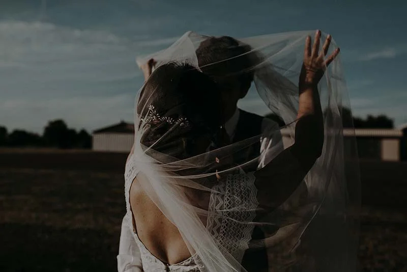 Jeunes mariés s'embrassant sous un voile blanc vaporeux dans une ambiance romantique et naturelle au coucher du soleil.