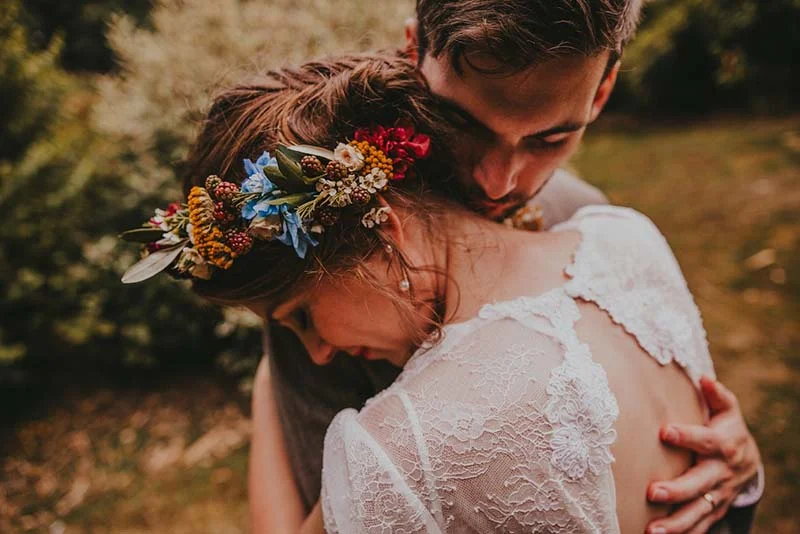 Couple enlacé dans un style champêtre, la mariée portant une couronne de fleurs colorées et une robe en dentelle blanche.