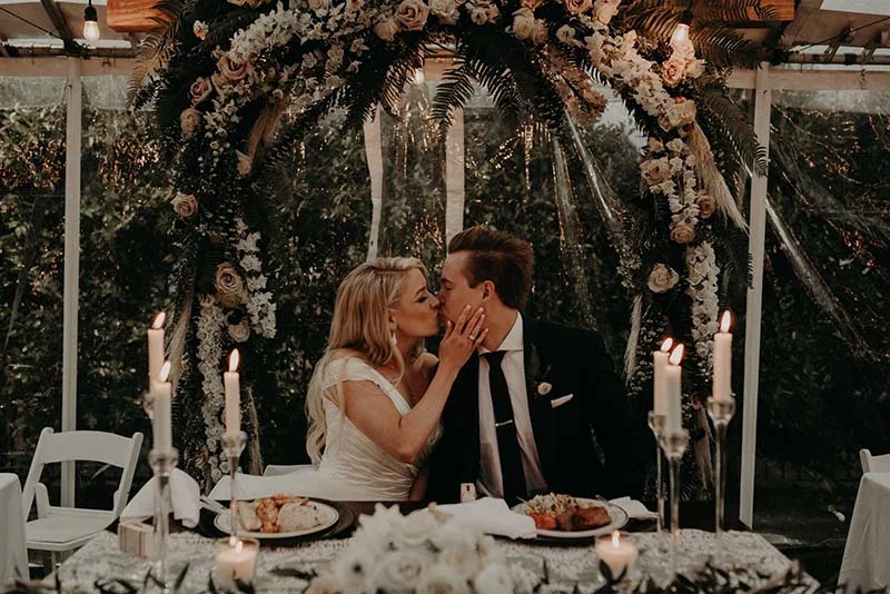 Jeunes mariés s'embrassant sous une arche florale romantique lors d'un dîner aux chandelles en extérieur.