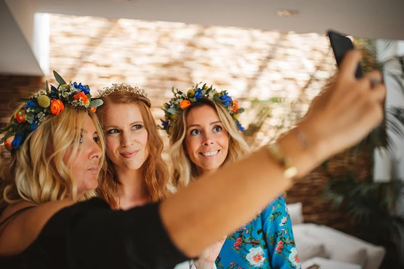 Trois femmes souriantes avec des couronnes de fleurs prennent un selfie dans une ambiance chaleureuse et bohème.