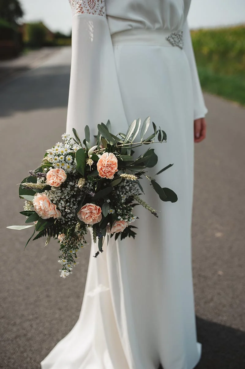 Mariée en robe blanche minimaliste tenant un bouquet champêtre de roses pêche et feuillage sur une route de campagne.