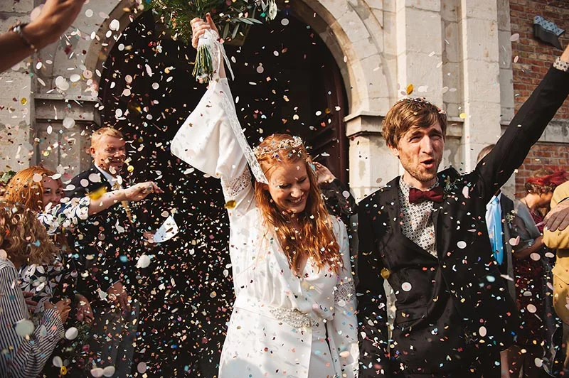 Jeunes mariés joyeux sous une pluie de confettis colorés, sortant d'une église dans une ambiance festive et bohème.