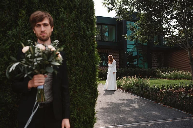 Un marié tenant un bouquet au premier plan, avec la mariée en robe blanche marchant vers une maison moderne en arrière-plan.