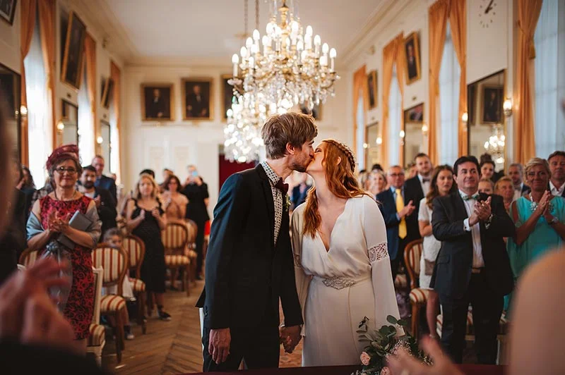 Un couple s'embrasse lors de sa cérémonie de mariage dans un salon classique, entouré d'invités et d'un grand lustre.