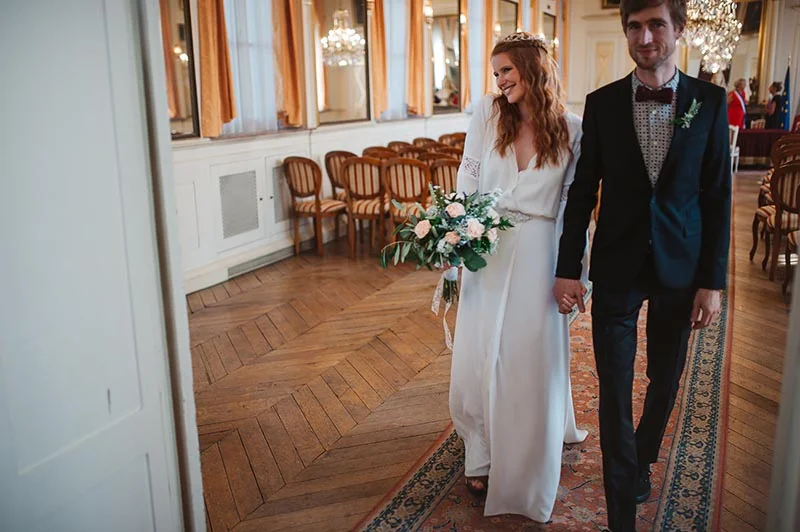 Jeunes mariés souriants marchant main dans la main dans un salon classique, elle en robe blanche et lui en costume noir.