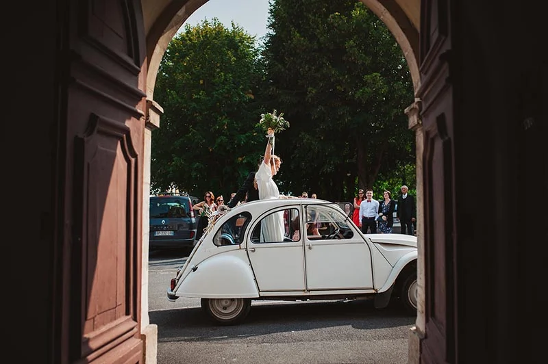 Une mariée brandit son bouquet depuis une Citroën 2CV blanche, entourée d'invités sous une arche en pierre.