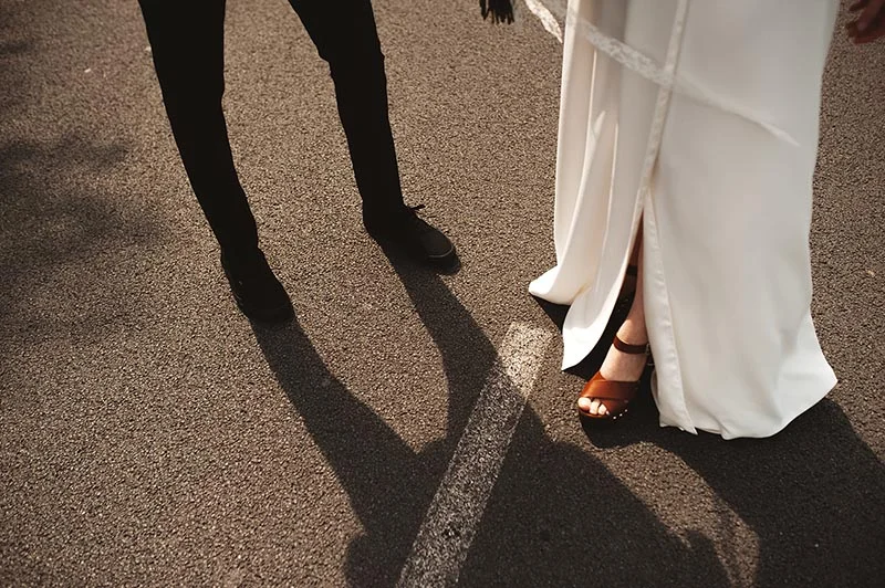 Gros plan sur les pieds d'un couple marié marchant sur le bitume, portant un costume noir et une robe blanche fluide.