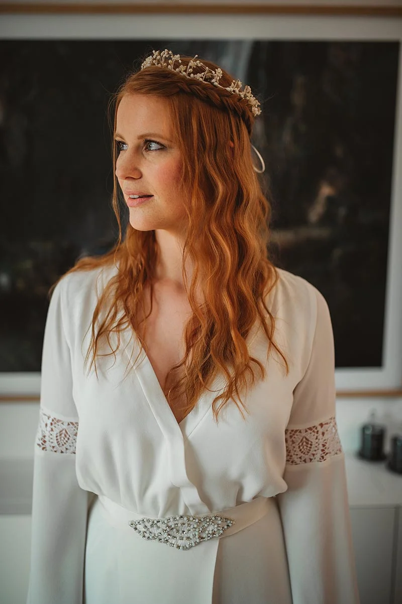 Future mariée aux cheveux roux ondulés portant un peigne doré et un peignoir blanc avec détails en dentelle.